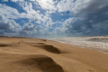 A vast sandy beach stretches towards a turbulent ocean under a dramatic sky filled with fluffy white clouds and patches of blue.