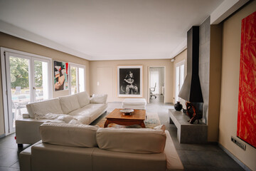Modern living room with white sofas, a wooden coffee table, a black fireplace, and wall art. Large windows bring in natural light.