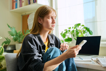 Serious teenage female sitting at home reading texting on smartphone