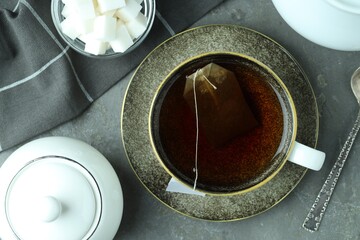 Tea bag in cup with hot drink, teapot, sugar cubes and spoon on gray textured table, flat lay