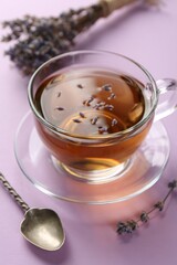 Aromatic lavender tea in glass cup, spoon and dry flowers on pink background, closeup