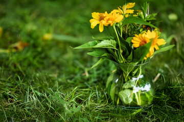 yellow and orange flowers in a glass vase on a wooden stand in tall green grass in a summer garden,...