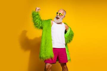 Confident senior man in eccentric green fur coat posing on yellow background, celebrating individuality and vibrant style.
