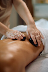 Close up of massage therapist using hot stones while massaging woman's back at health spa.
