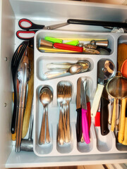 A cluttered desk drawer filled with various kitchen tools like forks, spoons, knives, and a whisk.