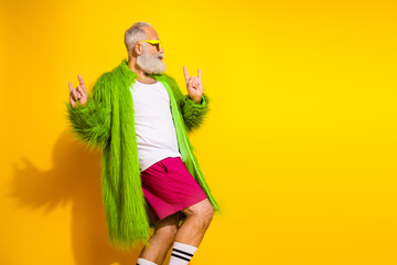 Stylish elderly gentleman in green fur coat and vibrant outfit posing against a yellow background...