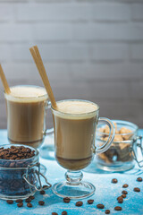 Two latte cups with eco friendly straws, coffee beans, and brown sugar on blue table