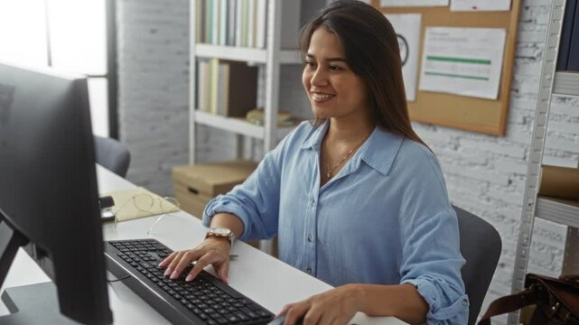 Woman working on a computer in a bright, organized office, showcasing her focus and professionalism with books and documents in the background making it a creative workplace setup.