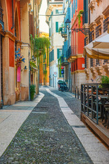 Obraz premium Italian narrow street with colorful building facades and cobblestone pavement in Verona old town, Veneto, Italy at sunrise. Medieval street with nobody. Vertical orientation. Travel destination