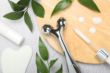Metal face roller, gua sha tool, cosmetic products and green leaves on gray background, flat lay