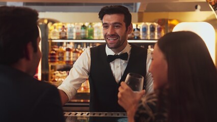 Focusing smiling smart bartender serving gin fizz or tonic soda at luxurious bar, engaging lively conversation with VIP customer couple at back view in atmosphere at lighting warm nightclub. Vinosity.