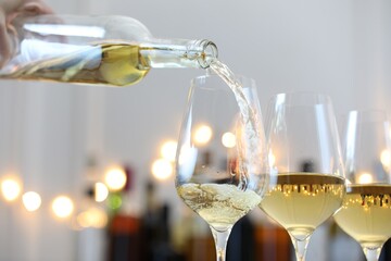 Pouring white wine into glass against blurred background with festive lights, closeup. Bokeh effect