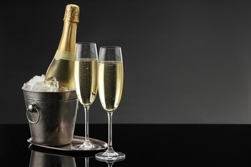 Bottle of sparkling wine in cooler bucket and glasses on black mirror surface. Space for text