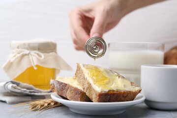 Woman pouring honey onto slices of bread with butter at grey table, closeup