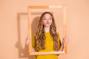 Young woman with wavy blonde hair posing playfully in a wooden frame with a minimalist beige...