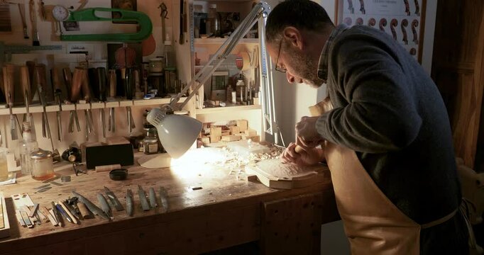 Slow motion shot of wood chips and shavings flying out as caucasian maker is carving belly bottom plate for a violin using gouge in workbench, tools in background, crafting a musical masterpiece