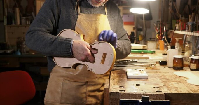 Close up of Luthier wearing an apron and gloves, carefully tampon ribs of a hand crafted violin in his workshop, illuminated by a lamp, surrounded by tools and materials, slow motion shot