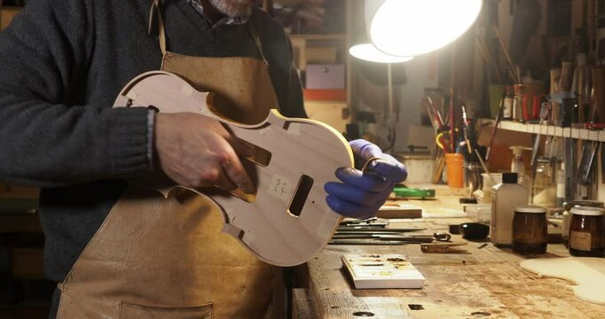 Close up of Luthier wearing an apron and gloves, carefully tampon ribs of a hand crafted violin in his workshop, illuminated by a lamp, surrounded by tools and materials, slow motion shot