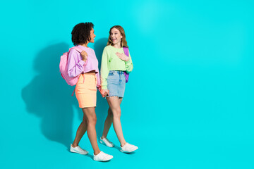 Two cheerful female friends walking and chatting on vivid teal background with trendy casual...