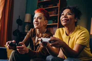 Smiling lesbians hold joysticks enjoying video games sitting on comfortable sofa
