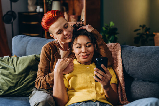 Woman shows pictures on smartphone to girlfriend sitting on sofa in living room
