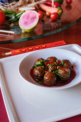 Vertical photo of glazed meatballs garnished with green onions and chili peppers, served in a white bowl on a decorative table. Chinese traditional food platter.