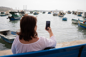 Fototapeta premium Woman traveler making photographs on a smartphone in fishing village Marsaxlokk on Malta
