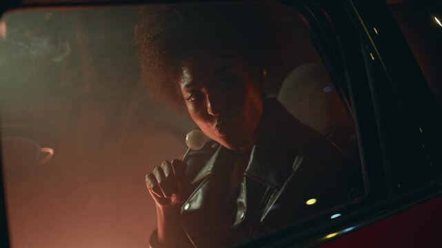Carefree girl licking lollipop sitting car backseat in night neon glow closeup.