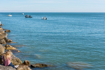 A woman with long blond hair is seen from behind sitting on a rock jetty looking out to sea where there  people fishing on boats,  Ponce Inlet, Florida, USA