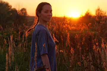A young woman stands in a sunlit field surrounded by plants during sunset. Her expression is serene...