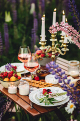 Elegant gorgeous wedding table decor or romantic dinner arrangement outdoors in blooming field. Purple lupine flowers, candles, fruits and wine, wooden vintage furniture. Sunset, summer, golden hour