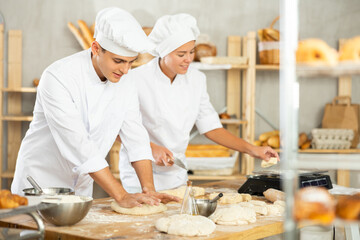 Friendly young guy processing dough while female baker weighing it by small portions in bakehouse