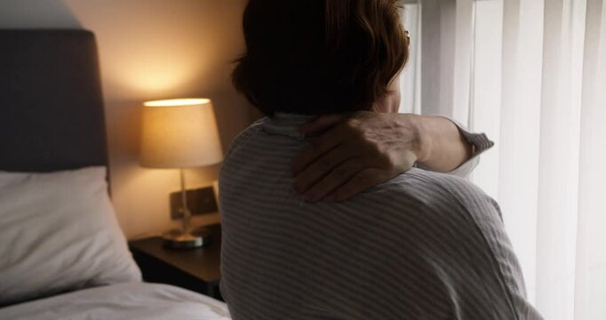 On the bed in a cozy bedroom, an elderly woman calmly holds her neck, feeling pain. The comfortable space around her can't conceal her suffering from the discomfort in her body.