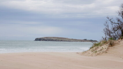 Windy beach with intense waves and sand movement under cloudy sky, featuring a distant rocky island and dry coastal vegetation © Víctor Sánchez