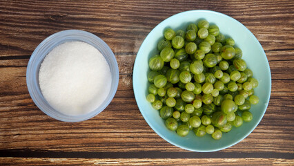 green gooseberries in blue plate next to plate filled with white sugar on brown wooden background. summer berry . top view
