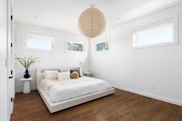 A minimalistic bedroom with dark hardwood flooring, a short bed with neutral bedding and pillows, and a circular light fixture hanging above.