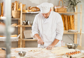 Professional baker in white uniform and cap cutting raw dough with knife to make croissants or buns