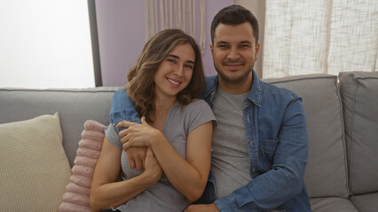 Obraz premium Man and woman couple sitting together on a couch in a cozy living room indoors, smiling and embracing each other warmly.