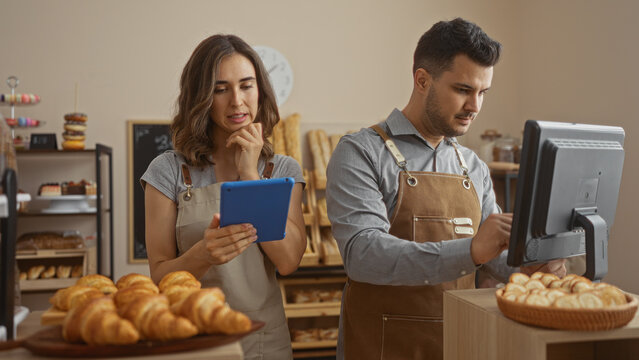 Woman and man working together in a cozy bakery interior wearing aprons while using digital tablet and computer with croissants and pastries on display