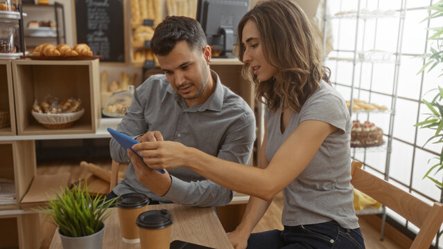 Man and woman sitting together in a bakery, looking at a tablet with coffee cups on the table - Powered by Adobe
