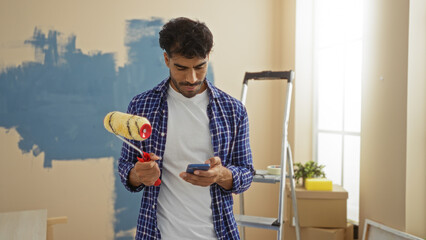 Young hispanic man painting room holding phone indoors suggesting home renovation in apartment with...