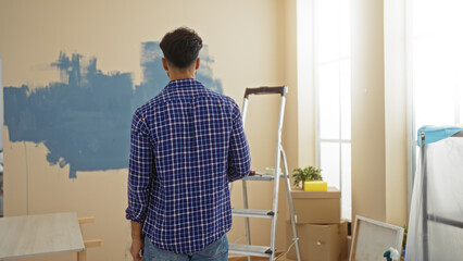 Man painting home interior with blue paint in a new apartment, standing in front of a wall, with a ladder and boxes around, suggesting home improvement and renovation.