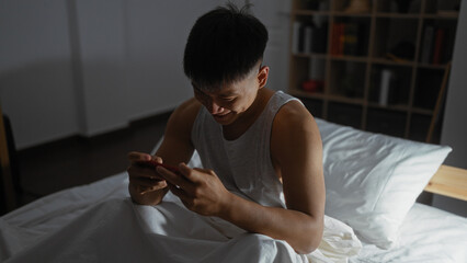 Young, chinese man using a phone while sitting on a bed in a dimly lit bedroom indicating a...