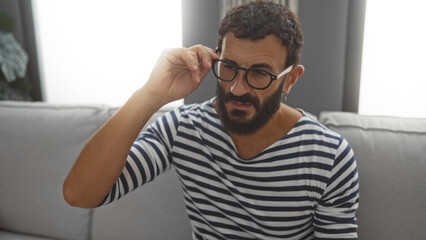 Obraz premium Young, hispanic, man with a beard adjusting his glasses while sitting in a living room of a modern home or apartment with a comfortable and stylish interior