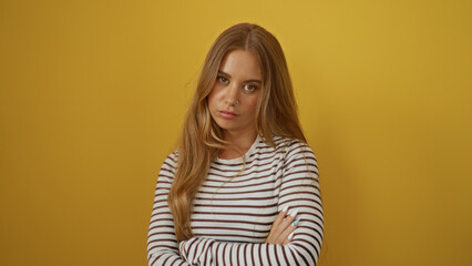 Obraz premium Blonde woman with crossed arms in striped shirt standing against an isolated yellow background looking pensive and beautiful.