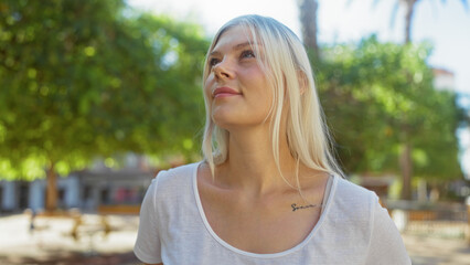 Young blonde woman in an urban park with green trees and a serene expression enjoying the outdoor...