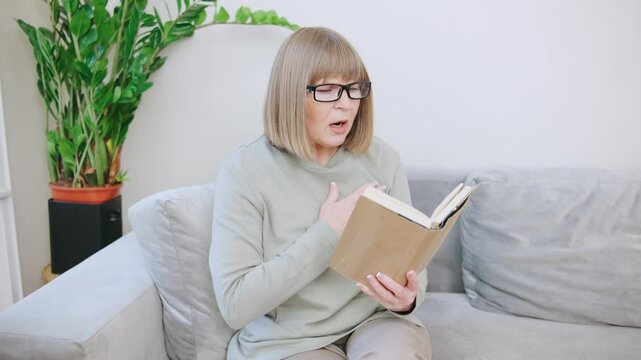 An elderly gray-haired woman with glasses is reading a book, sitting on the sofa in a cozy living room, her heart ached sharply. had a heart attack or stroke