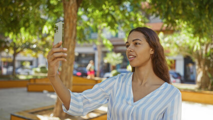 Naklejka premium Woman taking selfie outdoors in city park during daytime, brunette and beautiful in contemporary urban setting with trees and buildings.