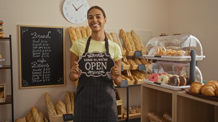 Young woman holding open sign in a bakery filled with bread and pastries, smiling and wearing an apron.