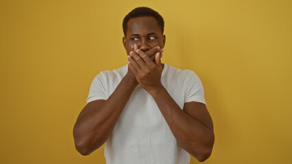 Young african man with hands on mouth standing against isolated yellow background, expressing surprise or shock, wearing casual white shirt, showcasing strong emotions.